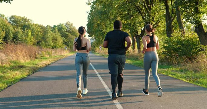 Back view of young mixed-races friends working out outdoor. Asian and Caucasian females doing cardio workout and running on road with African American man. Fitness training on nature. Sport concept