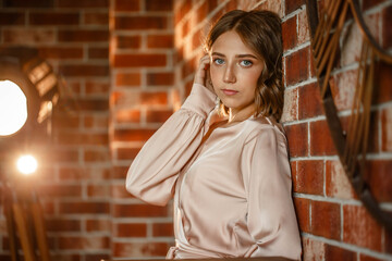  Beautiful young brunette in beige dress posing indoor near red brick wall. standing with his back against the wall