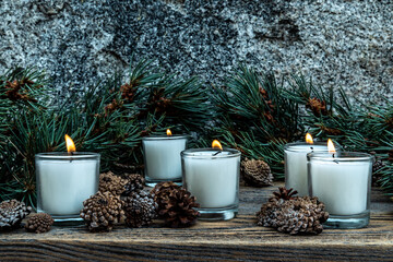 five lit white votive candles burning flames amid green pine branches and decorative pine cones