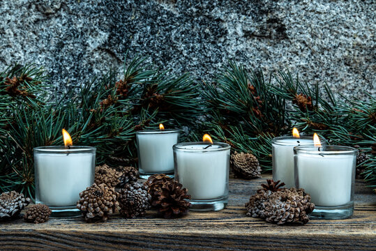 Five Lit White Votive Candles Burning Flames Amid Green Pine Branches And Decorative Pine Cones