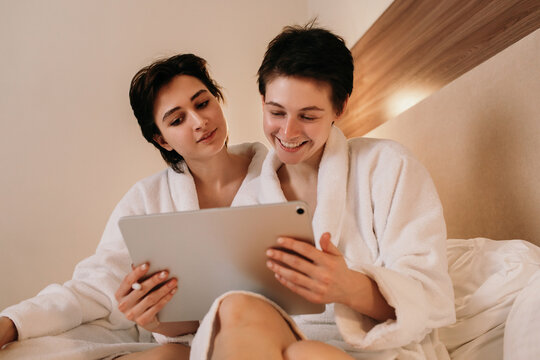Cheerful women in bathrobes using tablet on bed