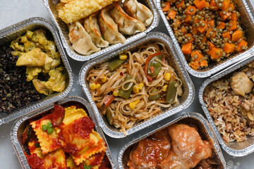 Food delivery. Different aluminium lunch box with ravioli, curry chicken  rice, gyoza tempura, noodles vegetables, lentils with pumpkin . airlines meals and snacks. takeaway takeout coronavirus food