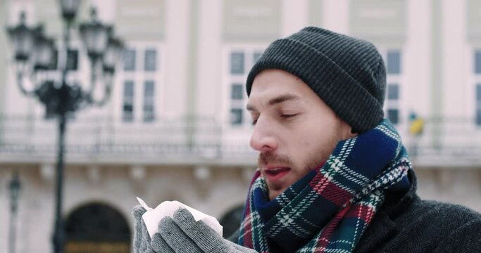 Portrait of Caucasian man in winter coatwith signs of cold sneezes into handkerchief on background of the city.