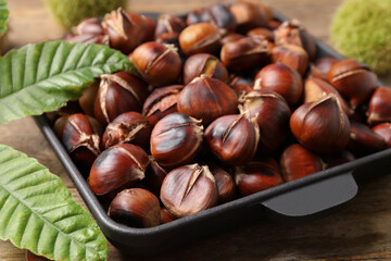 Delicious roasted edible chestnuts and green leaves on wooden table, closeup