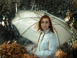 Woman with copper hair with umbrella and beige coat, walking in the forest