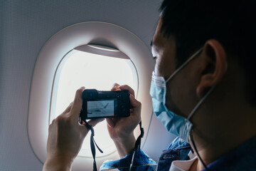 20s young adult Asian male tourist wearing a face mask inside aircraft cabin air and taking photos with a camera. Tourism during Covid-19 or Coronavirus pandemic. New normal travel concept