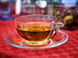 A transparent tea cup that is filled with tea and stands on a red tablecloth