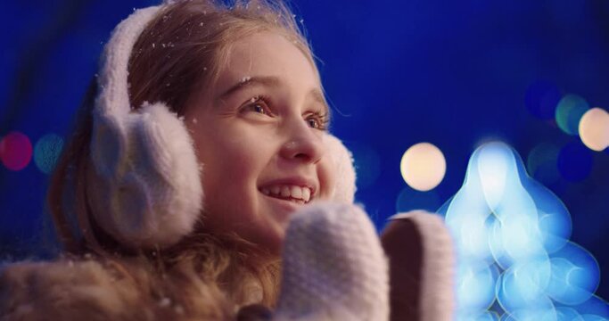 Close up portrait of cute little girl in winter clothes claps her hands while standing on the street. Schoolgirl watching New Year's show while it snows.