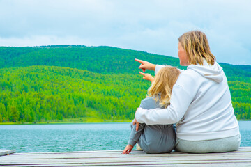 A woman and her child sit with their back to the camera with their hands raised against the backdrop of a mountain lake and mountains in the Altai Republic in Russia