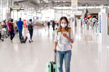 Young woman wearing a face mask and holding a mobile phone is looking for a check-in counter at the airport, New normal lifestyle concept