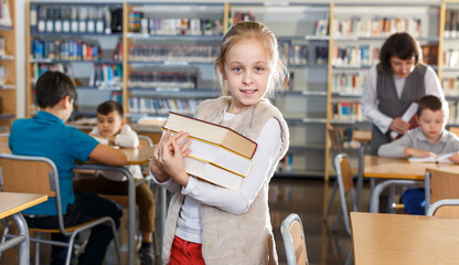 Portrait of happy cute preteen girl standing in classroom with books in hands ..