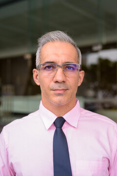Face of handsome Persian businessman with gray hair wearing eyeglasses outside modern building