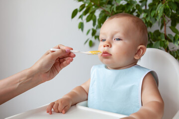 Mother feeds funny happy blond baby in blue bib from a spoon