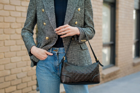 Young fashionable woman wearing gray tweed jacket with golden buttons, blue jeans, sweater and holding black leather baguette handbag. Street style.