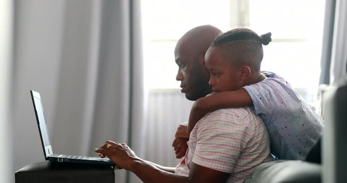 Black father working at home multi-tasking with child hugging
