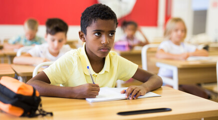 Diligent african american tween boy studying in classroom, listening to teacher and writing in workbook