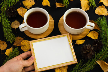 Holding emty photo frame near  two tea cups on black background with autumn leaves, pine branches and pine cones. Memories of past concept. Tea together. Copy space. Top view