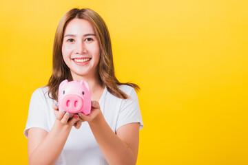 Fototapeta na wymiar Portrait Asian Thai beautiful happy young woman smiling holding pink piggy bank with a lot of money and looking to camera, studio shot isolated on yellow background, with copy space