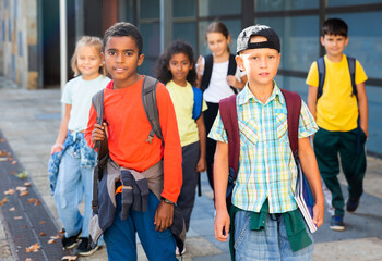 Multiethnic group of smiling tweenagers walking outside school building on autumn day, going to lessons.