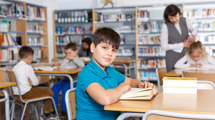 Fototapeta na wymiar Portrait of smiling intelligent preteen boy reading in school library on background with other students and teacher/