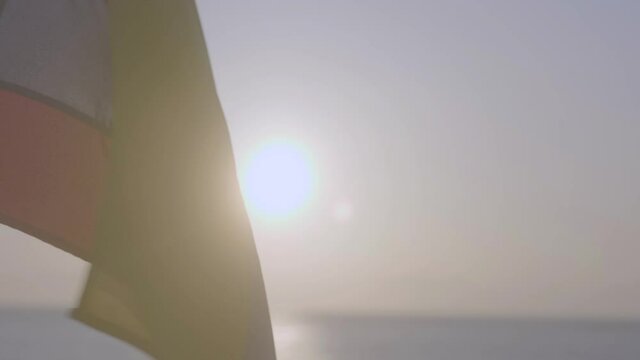 A flag of Colombia or Ecuador is being moved and flamed by the wind. The sun is setting on the horizon, the sunlight is being reflected on the ocean.