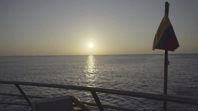 The sun is setting on the horizon and the sunlight is being reflected on the water of the ocean while a flag of Ecuador or Colombia flames near a railing.
