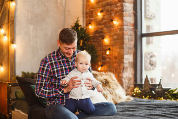 Little boy with his father in Christmas loft bedroom interior. Christmas celebration. Boy with Dad weared in casual. New year eve in loft room with red accents. Lighting vintage garland on background