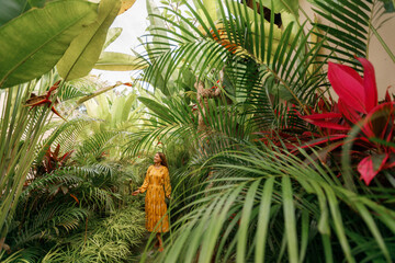 Ypoung happy woman walking in tropical garden in long yellow dress. Brunette with long dark hair ...