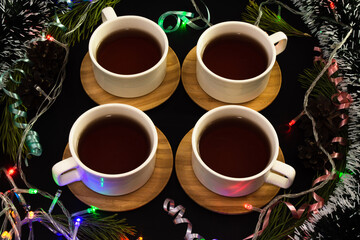 Four tea cups surrounded by tinsel, luminous garland, colourful curly ribbons and pine branches on black background. Celebrating christmas in company concept