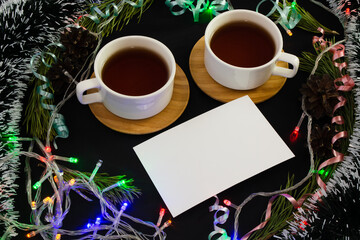 Two tea cups and blank white sheet, photo card surrounded by luminous garland, tinsel, pine branches and curly colourful ribbons on black background. Celebrating Christmas together concept. Copy space