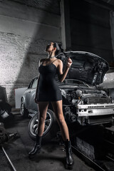 Naklejka na ściany i meble Portrait woman in dress posing next to a car in the garage, in background old car. Creative Colorful Bright Portrait .Design Car workshop art concept