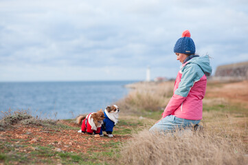 pensive healthy sport young woman in hat and track suit sitting before two dressed small pets chihuahua dogs on cold weather spring sea shore meditating