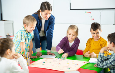 Group of school kids with teacher sitting together around desk in classroom, playing educational tabletop game