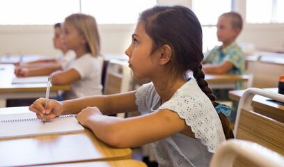 Diligent elementary school student tween girl studying with classmates, making notes of teacher...