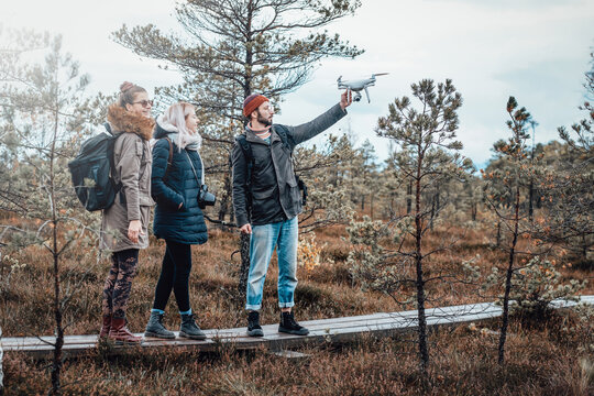 Bearded guy showing quadcopter holding it with raised hand and two girls they stay behind guy in autumn relaxed forest.