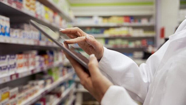 Close up of female pharmacist scrolling on digital tablet checking and counting stock of medicines on shelves in pharmacy 