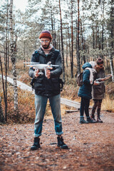Bearded guy stays holding quadcopter and preparing it behind his friends in autumn lovely forest.