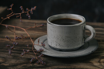 A cup of espresso coffee on a wooden table on a dark background. A sprig of dried flowers nearby.