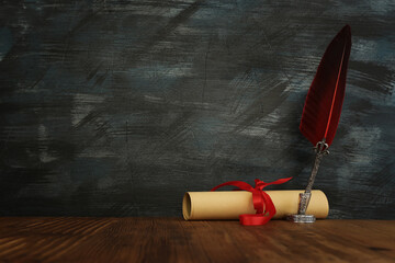 Old feather quill ink pen with inkwell and paper scroll over wooden desk in front of black wall...