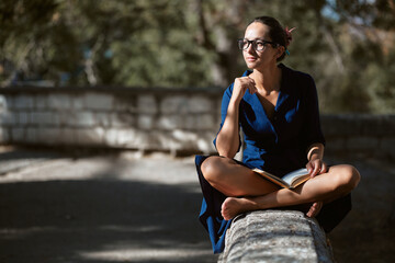 Pensive young woman in glasses looking away sitting in park. Street style, read book and city walk concept, copy space