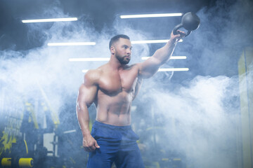 Portrait of a muscular sportsman performing kettlebell swings with one hand in a gym standing in...