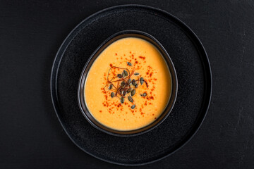 Homemade pumpkin soup with pumpkin seeds, chili flakes, pink pepper in black bowl on black slate background. Vegan soup. Top view.