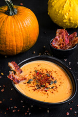 Homemade pumpkin soup with pumpkin seeds, chili flakes, pink pepper, fried bacon and some types of winter squash in black bowl on black slate background. Vegan soup. 