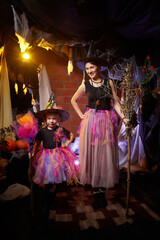 Beautiful brunette mother and cute little daughter looking as witches in special dresses and hats in room decorated for Halloween. Witchcraft and wizardry in carnival. Halloween style photo shoot.