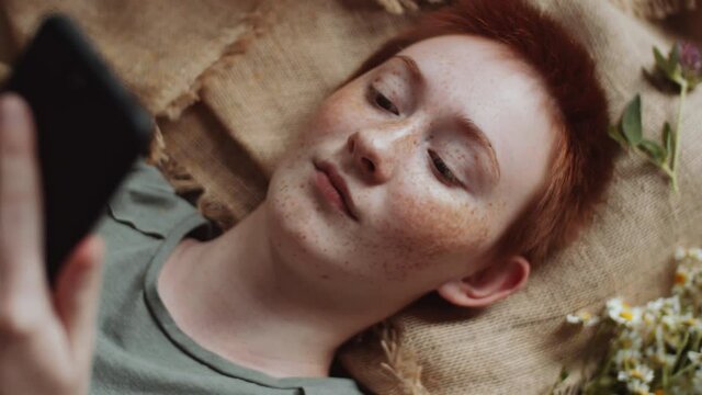 Top view close up shot of young pretty redheaded woman lying on burlap fabric with wildflowers and using smartphone