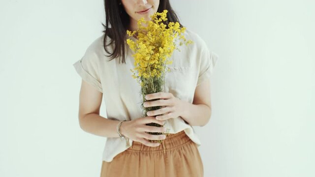 Midsection studio shot of young brunette woman standing against white background, holding bouquet of yellow flowers and enjoying scent
