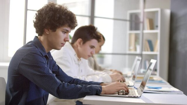 Slow-mo side-view medium closeup of team of young programmers sitting in row at desk typing on laptops with concentration
