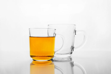 Two transparent glass mugs, one is empty and other with yellow liquid, tea or juice, on white reflective surface.