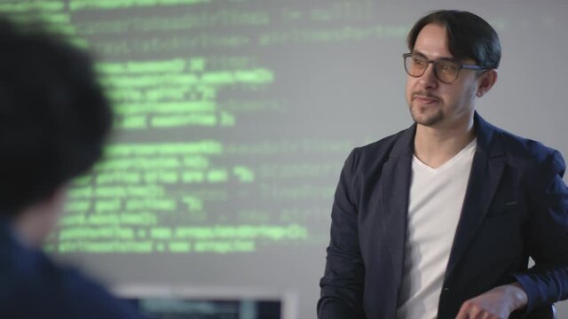 Panning footage of back-view row of teenagers listening to young male computer programming teacher explaining new topic with projector full of codes at background