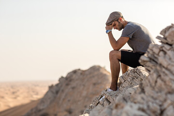 Depressed young man feeling sad sitting alone on top of a mountain cliff overlooking the desert.